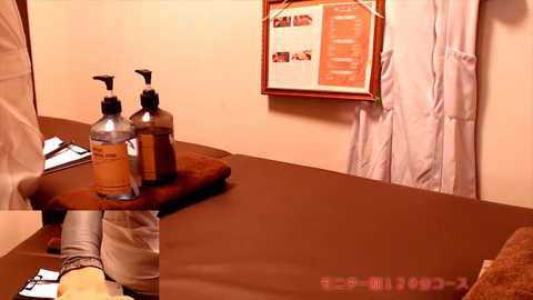 Media: Video of a dimly-lit, minimalist Japanese-style room with brown walls and tatami mats. A wooden tray holds two brown bottles of soap, a towel, and a clipboard. A white apron hangs on the wall.