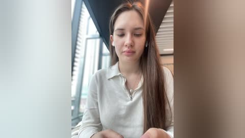 Media: Video of a young Caucasian woman with long, straight brown hair, wearing a white collared shirt, standing indoors near large windows with blinds. She has a calm expression, looking downward.