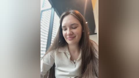 Media: Video of a young woman with long brown hair, wearing a light-colored shirt, smiling, standing near a window with partially open white shutters, in a bright, modern interior.