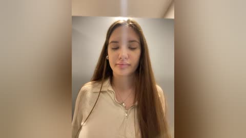 Media: Video of a young woman with long, straight brown hair, wearing a beige cardigan, standing in a dimly lit hallway with blurred beige walls.