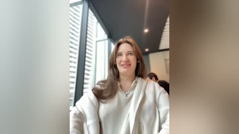 Media: Video of a smiling Caucasian woman with long brown hair, wearing a cream-colored sweater, standing indoors near large windows with white blinds and a modern ceiling.