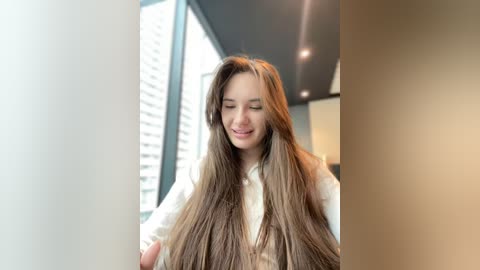 Media: Video of a young woman with long, straight, brown hair, smiling, standing in a modern, bright room with large windows and tall buildings in the background. She wears a white shirt.