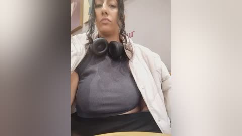 Media: Video of a woman with curly hair, wearing a gray shirt, black skirt, and white jacket. She's seated indoors with headphones around her neck.