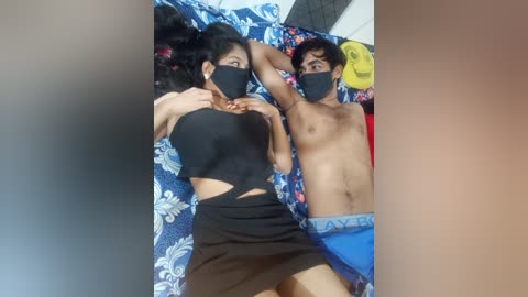 Media: Video of two shirtless young men, one with a black mask and black crop top, lying on a bed with blue floral sheets. The other man, shirtless, is partially visible in the background.