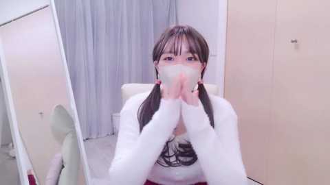 Media: A video of an Asian woman with long black hair in pigtails, wearing a white sweatshirt and face mask, sitting in a white chair, hands clasped in front. The background features beige walls and a white mirror.