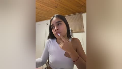 Media: Video of a young woman with long dark hair, fair skin, and slender physique, wearing a white one-shoulder top, standing in a room with wooden ceiling and beige walls. She touches her lips with her fingers.