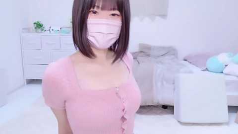 Media: A video of an Asian woman with straight black hair and a pink face mask, wearing a pink ribbed top, standing in a minimalist, white-themed bedroom with a bed and dresser.