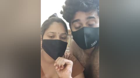 Media: Video of a South Asian couple with medium skin tones, wearing black masks, holding hands, in a dimly lit room.