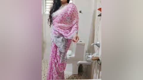 Media: Video of a woman in a pink and white floral saree standing in a bathroom with a white toilet, beige tiled floor, and a window with white blinds.