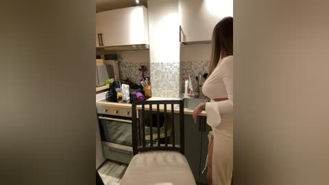 Media: Video of a modern, cozy kitchen with white cabinets and a woman in a white robe standing near a wooden chair.