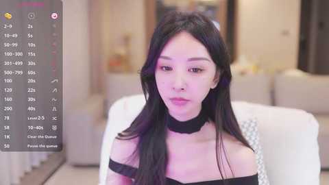 Media: Video of an East Asian woman with long black hair and fair skin, wearing a black off-shoulder top, sitting on a white couch in a modern living room.