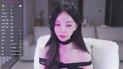 Media: Video of an East Asian woman with long black hair, wearing a black choker, seated in a modern living room with a blurred background.