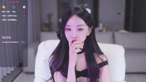 Media: Video of an East Asian woman with long black hair, wearing a black choker and bra, seductively biting her finger, in a modern, dimly lit living room.