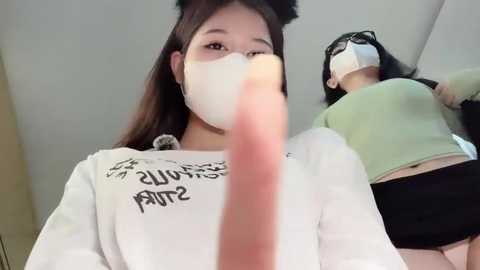 Media: Video of two Asian women in casual outfits, one with a white mask, the other wearing a green top and black shorts. Background shows a beige wall and a slanted ceiling.