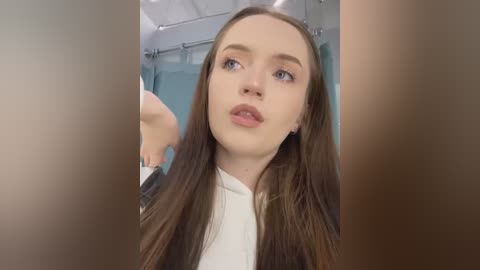 Media: Video of a young Caucasian woman with long brown hair, light skin, and blue eyes, wearing a white top, standing in a modern bathroom with glass shower and light-colored tiles.