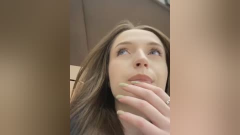 Media: Video of a young woman with fair skin, blue eyes, and long brown hair, standing in a beige hallway, looking up with a contemplative expression. Her hand is near her mouth, fingers touching her lips.