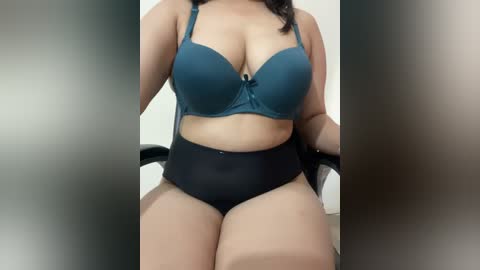 Media: Video of a plus-size woman with dark skin, wearing a teal bra and black high-waisted panties, sitting in a black chair, with blurred background.