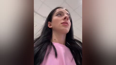 Media: Video of a young woman with long black hair, wearing a pink top, gazing upwards, with a ceiling of white tiles in the background.