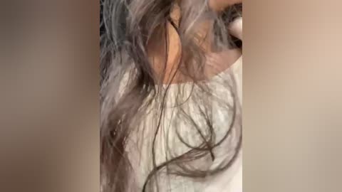 Media: Video of a woman's partially visible face, with long, wavy, dark brown hair cascading over her shoulders. Her skin tone is light brown, and she appears to be wearing a light-colored, possibly white, top. The background is blurred, focusing attention on her hair and face.