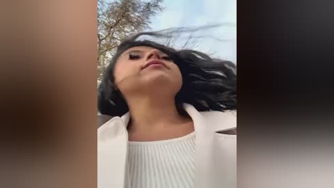 Media: Video of a young woman with long black hair, fair skin, and a confident expression, wearing a white sweater, standing outdoors against a cloudy sky and leafy tree.