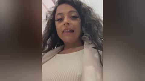 Media: Video of a Black woman with curly hair, wearing a white ribbed top, and beige cardigan, smiling with a slight open mouth, taken indoors.