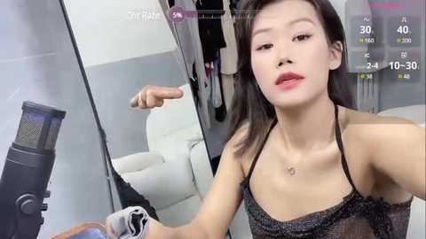 Media: Video of a young East Asian woman with fair skin, straight dark hair, wearing a black lace dress, sitting in a modern, minimalist room with a white chair and mirror. She's holding a black hair dryer.