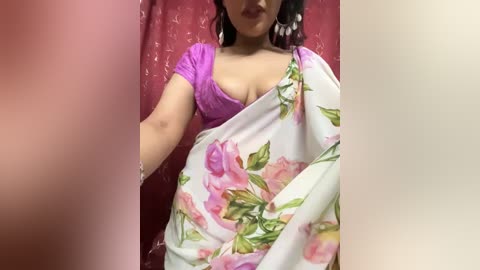 Media: Video of a woman with medium skin tone, wearing a purple top and floral saree, partially covering her chest, against a pink, textured curtain background.