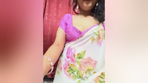 Media: A video of a smiling woman in a pink saree with floral motifs, wearing a purple blouse and large earrings, standing against a red backdrop.