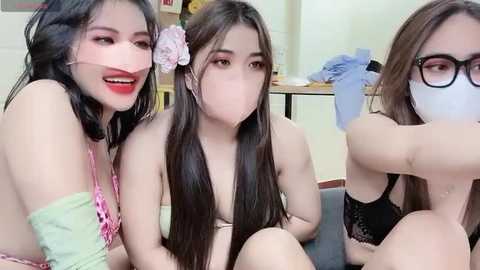 Media: Video of three young Asian women, wearing face masks, sitting closely together, with one wearing a pink flower in her hair, on a sofa.