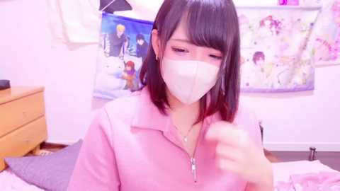 Media: Video of a young Asian woman with straight black hair, wearing a pink polo shirt and white mask, seated indoors, with a white wall, floral poster, and wooden dresser in the background.