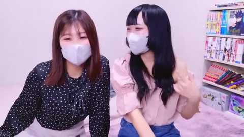 Media: Video of two Asian women wearing face masks, one in a black polka-dot blouse, the other in a pink blouse, seated on a bed in a brightly lit room with shelves of anime figurines in the background.