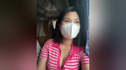 Media: Video of a woman with light skin, wearing a white face mask, pink and white striped polo shirt, and a blue jacket, seated in a bus.