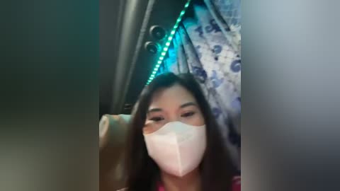 Media: Video of a woman with long dark hair, wearing a white mask, sitting in a dimly lit room with a patterned curtain and blue LED lights.