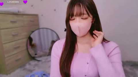 Media: Video of a young Asian woman with long brown hair, wearing a light pink face mask and long-sleeved sweater, standing in a cluttered, beige room with a mirror and a dresser.