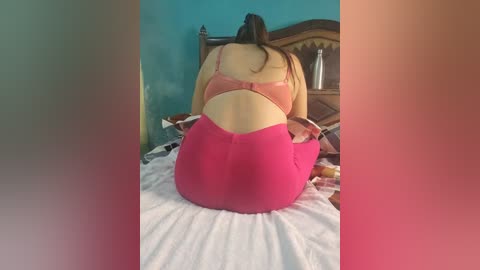 Media: Video of a woman kneeling on a bed, wearing a pink bra and high-waisted pink pants. She has long, straight dark hair. The background shows a wooden headboard and a teal wall.