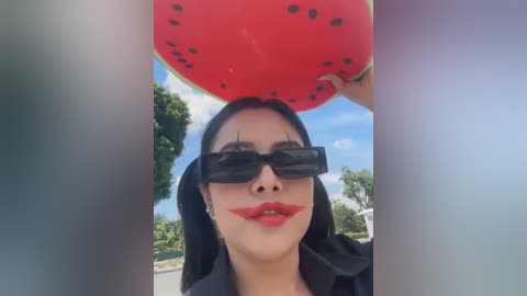 Media: A video of a person with dark hair, wearing black sunglasses and red clown makeup, holding a large watermelon slice overhead against a bright blue sky and green foliage.