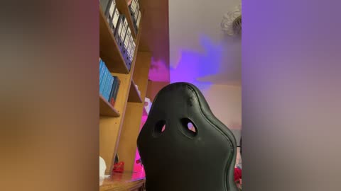 Media: Video of a person wearing a black, eyeless mask sitting in a wooden bookshelf room with purple and blue lighting.