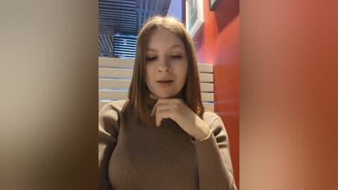 Media: Video of a young Caucasian woman with light skin and shoulder-length brown hair, wearing a brown sweater. She stands in a narrow, brightly lit hallway with orange walls and horizontal blinds.