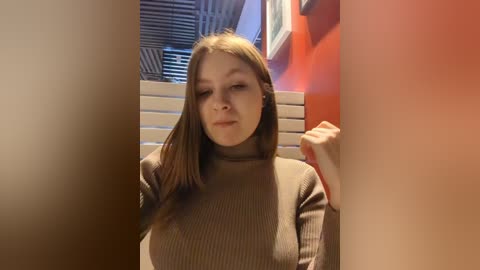 Media: A video of a young woman with fair skin, long brown hair, and a neutral expression, wearing a beige ribbed turtleneck sweater, standing in a room with orange walls, a window with blinds, and white shelving units.