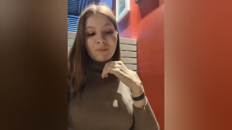 Media: Video of a young Caucasian woman with straight, light brown hair, wearing a dark green turtleneck sweater, standing in a bright red room with white blinds and a white radiator.