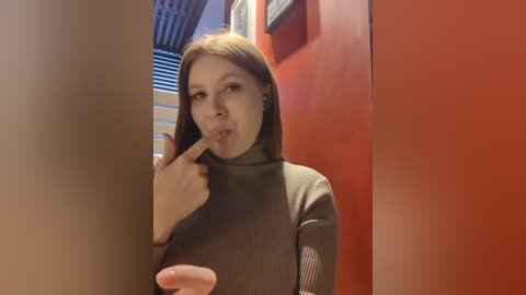 Media: Video of a young woman with straight, light brown hair, wearing a green turtleneck sweater, standing in front of a red wall, making a \"shh\" gesture with her finger to her mouth.