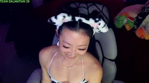 Media: A video of an Asian woman with fair skin, wearing a cow print headband, a white bikini, and a gold necklace, seated on a gaming chair in a dimly lit room.