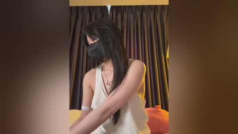 Media: Video of a woman with long black hair, wearing a black face mask and a beige sleeveless top, standing in front of dark curtains in a dimly lit room.