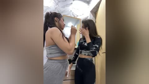 Media: A video shows two women in a bathroom; one with long dark hair in a grey crop top and leggings, the other with long brown hair in a black and white patterned top and leggings.