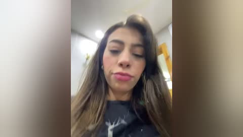 Media: Video of a young woman with long brown hair, light skin, and closed eyes, making a pouty face while wearing a black T-shirt with a white deer graphic. Indoor setting with blurred background.