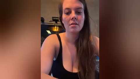 Media: Video of a white woman with long brown hair, wearing a black tank top, sitting in a dimly lit room with kitchen appliances in the background.