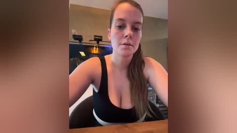 Media: A video of a fair-skinned, fit woman with long brown hair in a ponytail, wearing a black sports bra and black leggings, leaning forward on a table in a dimly lit kitchen.