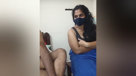 Media: Video of a South Asian woman with medium skin tone and curly black hair, wearing a blue mask and black tank top, sitting on a toilet, legs crossed, in a bathroom with white walls.