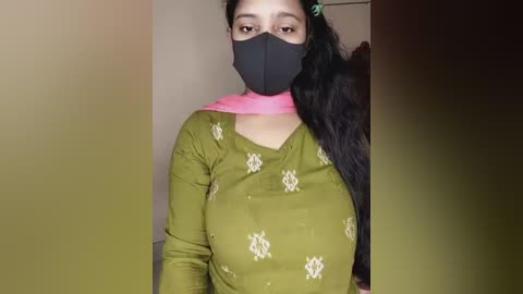 Media: A video of a young woman with dark skin, wearing a black mask, green top with floral embroidery, and a pink scarf, standing indoors against a beige wall.