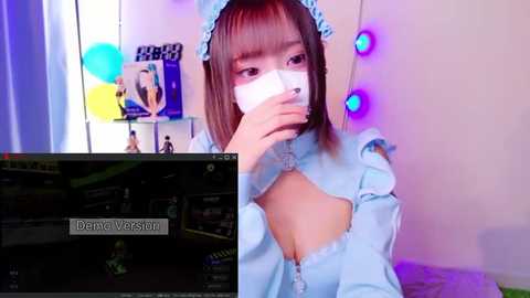 Media: A video of an East Asian woman in a light blue maid costume, with a mask covering her nose and mouth, sitting indoors. The background features a TV screen displaying the game \"Demonslayer.\